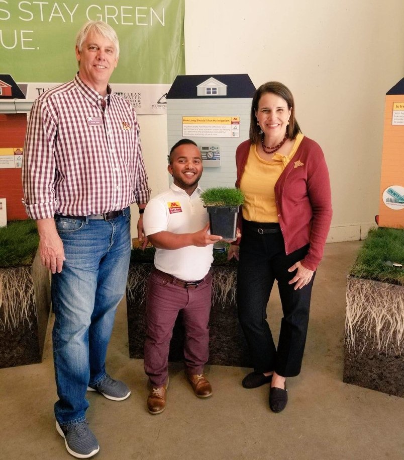 Three people stand in front of the turfgrass state fair display with a pot of fine fescue turfgrass